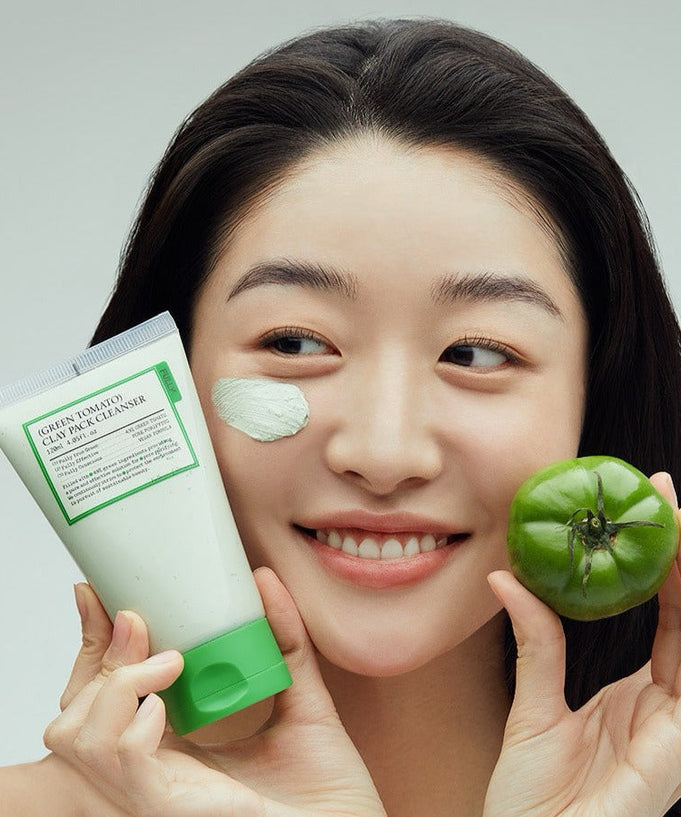 Woman holding FULLY Green Tomato Clay Pack Cleanser 120ml with a green tomato, smiling with clay on cheek.