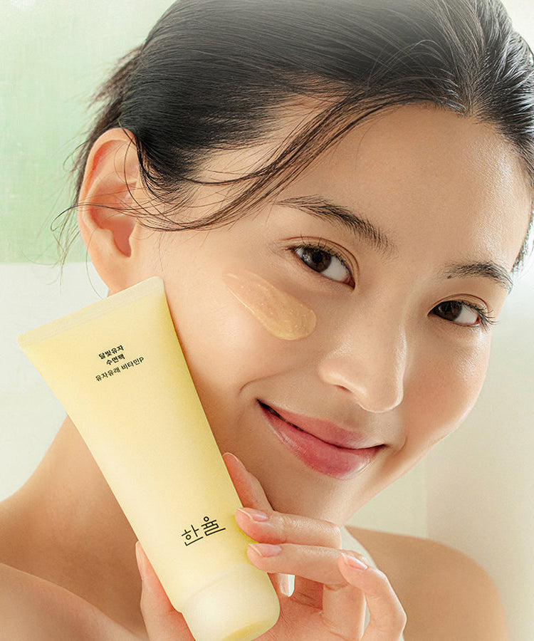 Woman applying HANYUL Moonlight Yuja Sleeping Pack 100ml on cheek, showcasing the tube and texture of the skincare product.