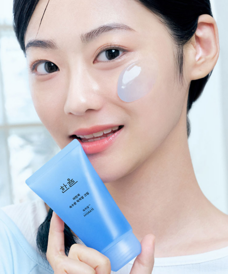 Smiling person holding HANYUL Young Artemisia Extract Moisturizing Cream 60ml, applying cream on cheek.