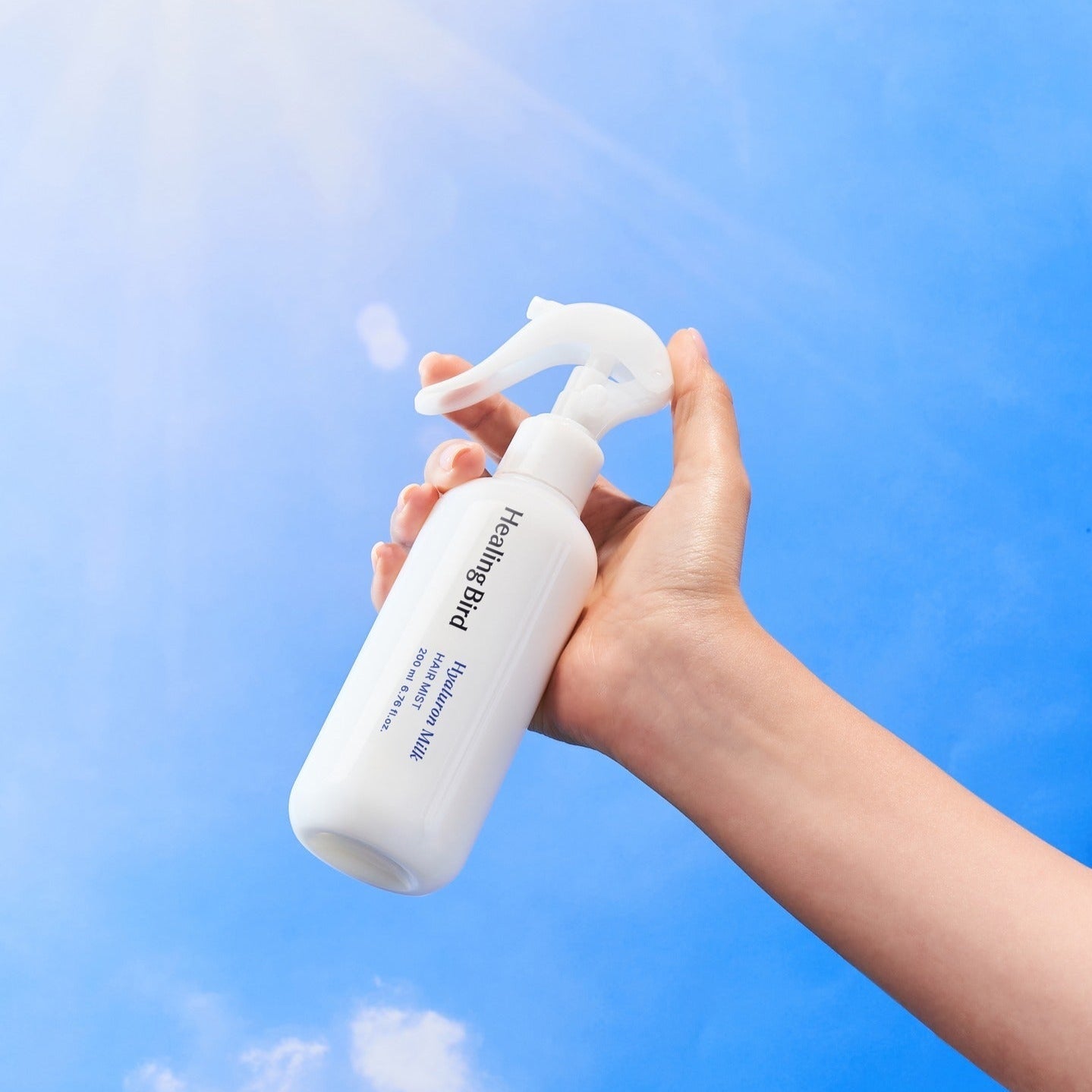 Hand holding HEALING BIRD Hyalutonic Milk Hair Mist 200ml against blue sky background.