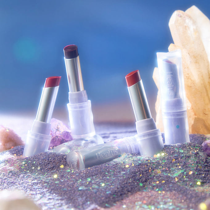 Assorted HEALUS Bare Lip Glow 4g lipsticks displayed on glittering sand next to crystals.