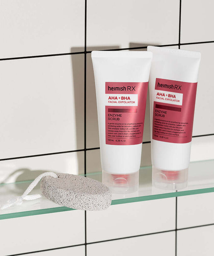 HEIMISH RX AHA BHA Enzyme Scrub 130ml displayed on a glass shelf with a pumice stone in a tiled bathroom setting