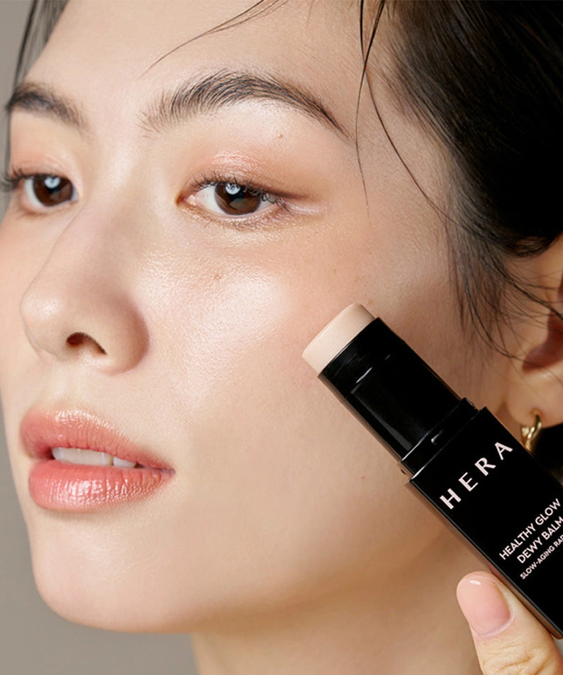 Woman applying HERA Healthy Glow Dewy Balm 10g to her cheek for a radiant look.