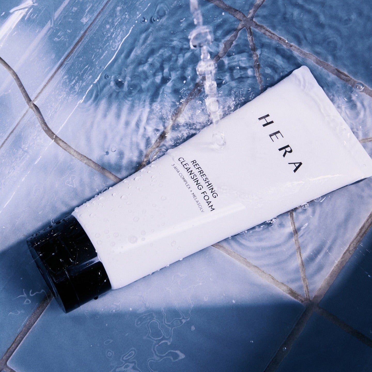 HERA Refreshing Cleansing Foam 160g tube under running water on a tiled surface, showcasing its refreshing cleansing properties.