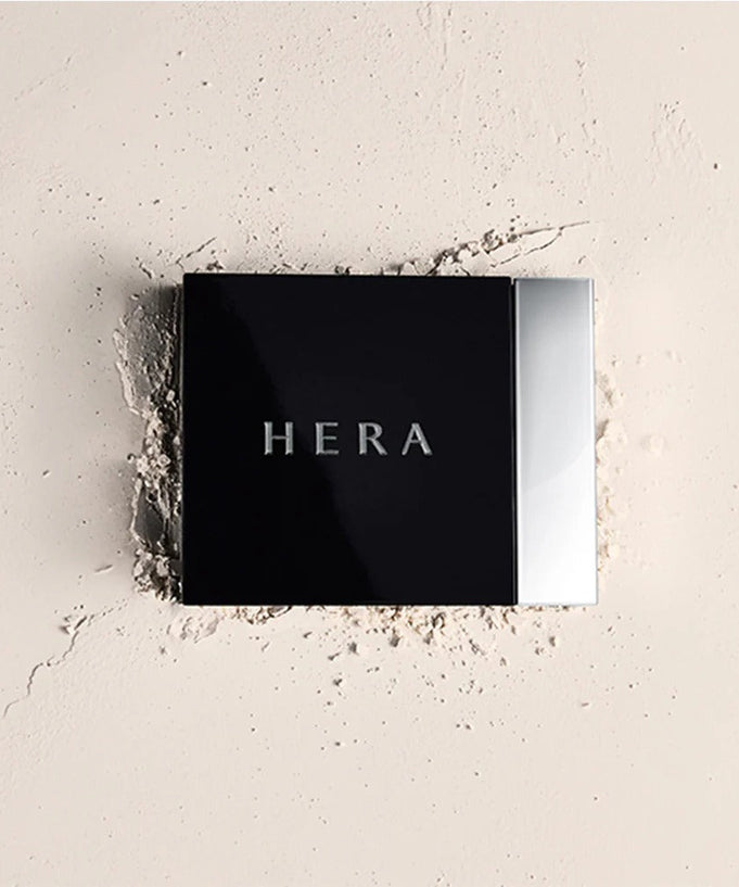 Close-up of HERA Airy Powder Primer 8.5g on a textured surface, showcasing sleek packaging and brand name.