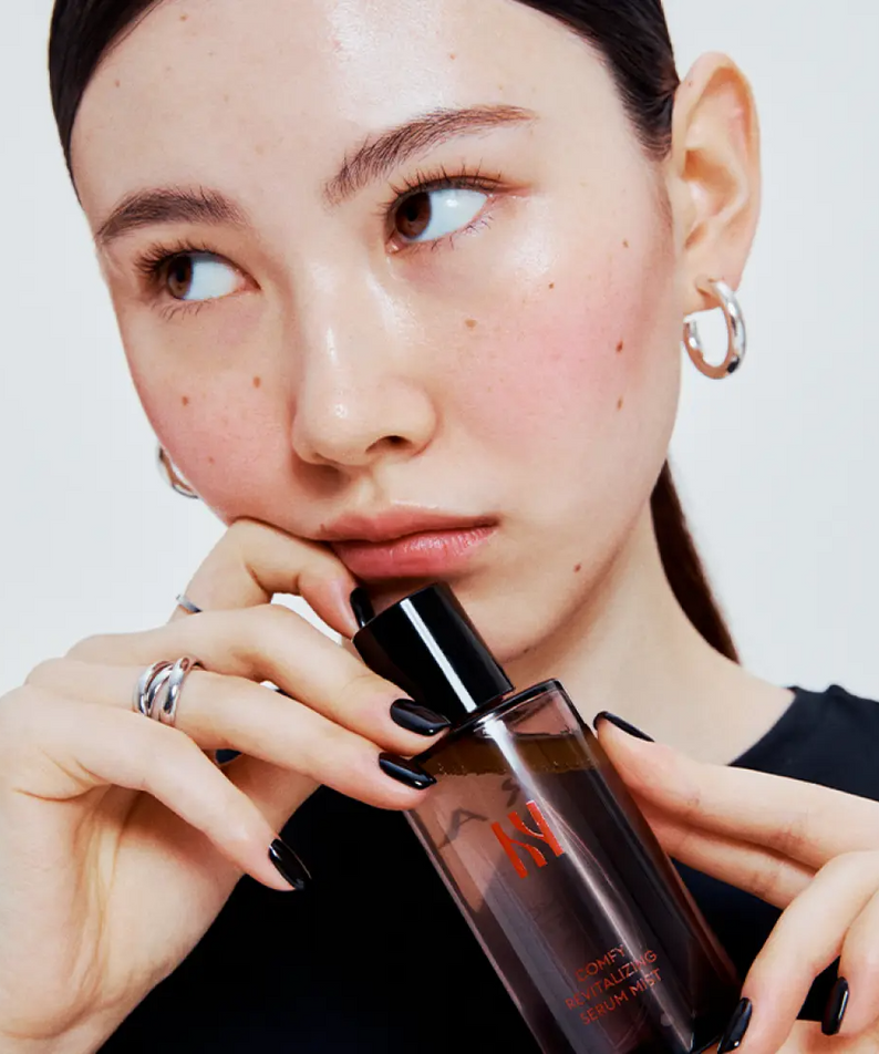 Woman holding HERA Comfy Revitalizing Serum Mist 90ml bottle, showcasing skincare product for healthy complexion.