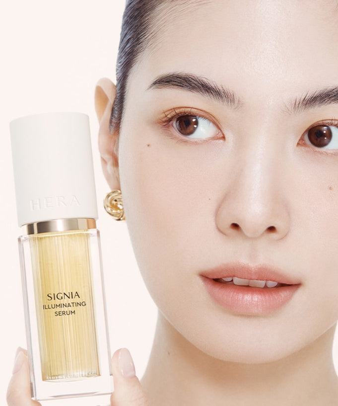 Woman holding HERA Signia Illuminating Serum 30ml with close-up facial expression.