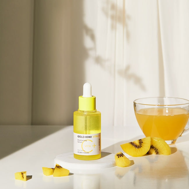 HOLIKA HOLIKA Gold Kiwi Vita C+ Brightening Serum 45ml bottle with sliced kiwi and a glass of juice on a sunlit table.
