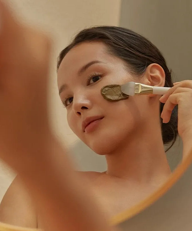 Woman applying HOLITUAL Clean Therapy Powder Mask with brush in mirror reflection
