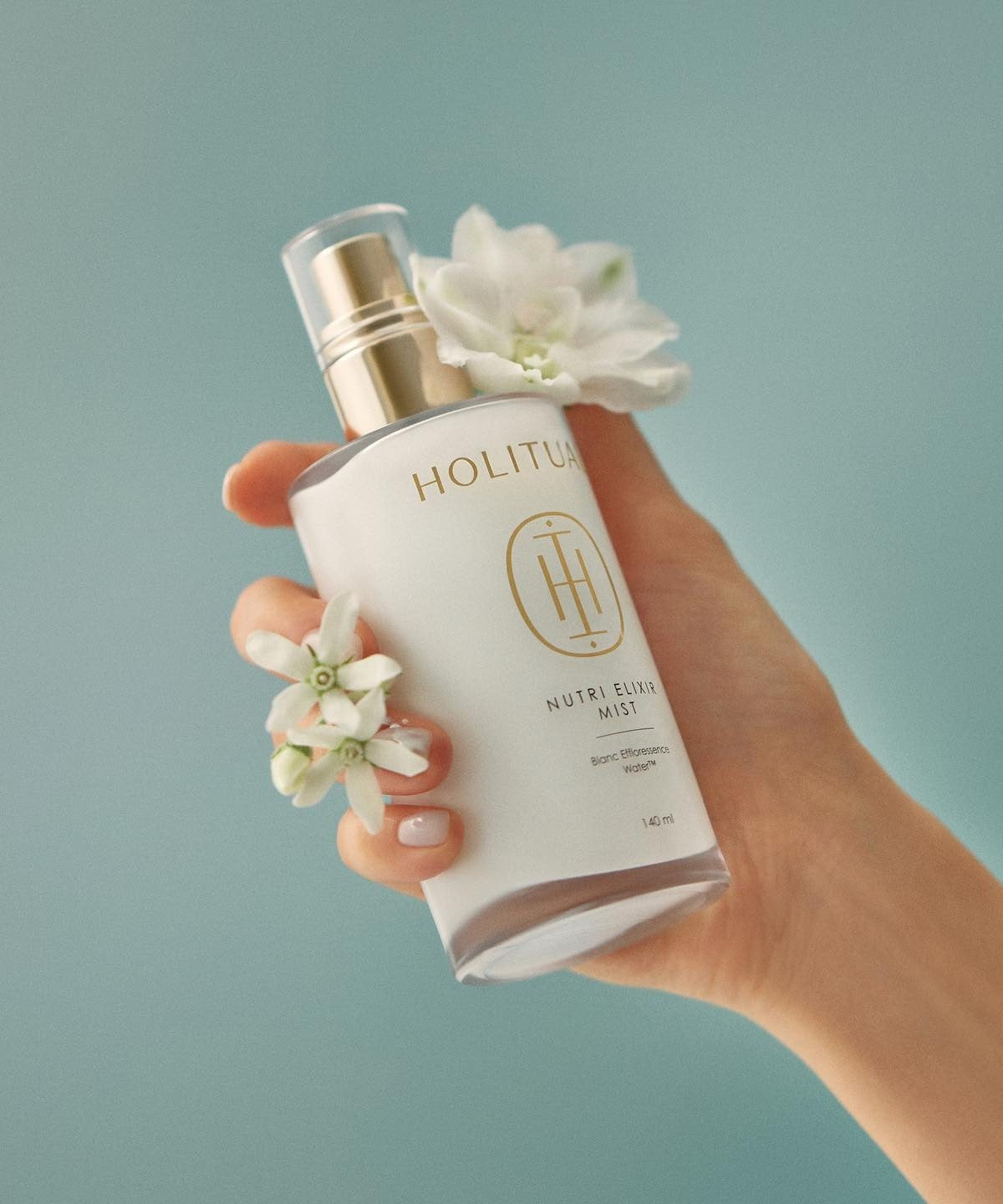 Hand holding HOLITUAL Elixir Mist 140ml bottle with flowers accentuating the design.