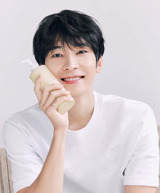 Person holding a bottle of HUXLEY Body Lotion Blue Madina Tangerine, smiling in a white t-shirt.