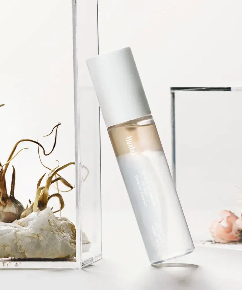 Bottle of HUXLEY Oil Mist Sense Of Balance next to decorative plants on a white background.