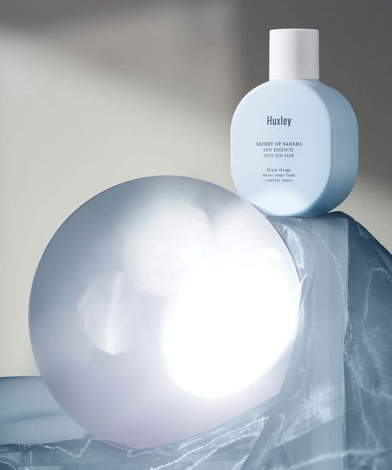 HUXLEY Sun Essence Stay Sun Safe product displayed with artistic lighting and draped fabric backdrop.