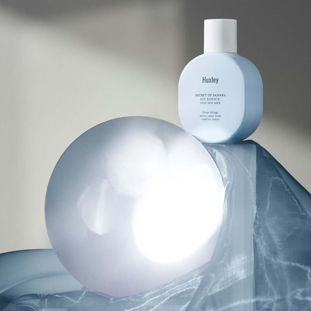 HUXLEY Sun Essence Stay Sun Safe product displayed with artistic lighting and draped fabric backdrop.
