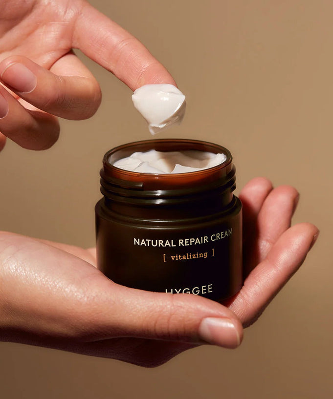 Close-up of hands holding HYGGEE Nature Repair Cream 50ml jar with cream on fingertip, showcasing its texture and packaging.