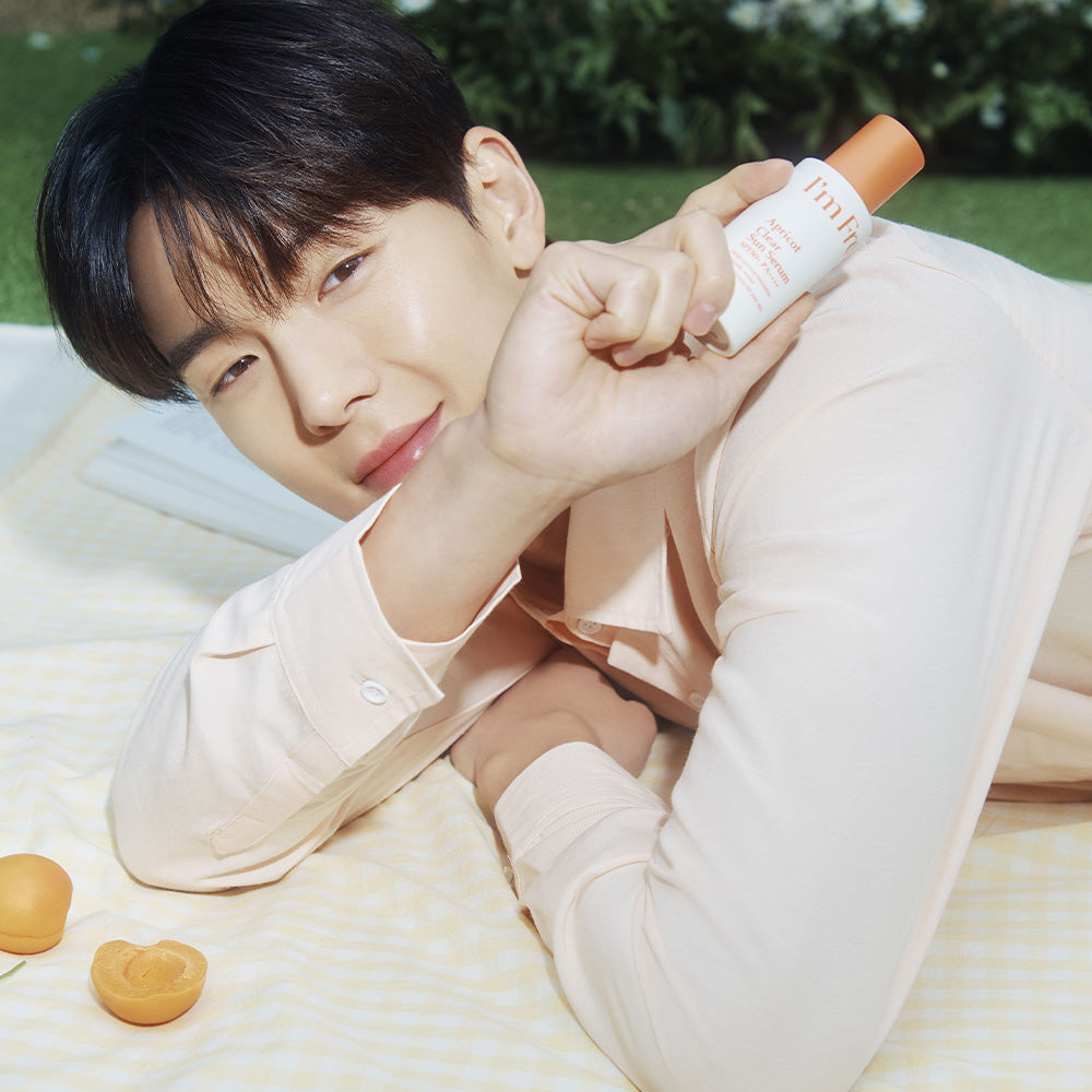 Person holding I'M FROM Apricot Clear Sun Serum 50ml outdoors on pastel blanket with apricots; skincare product displayed.