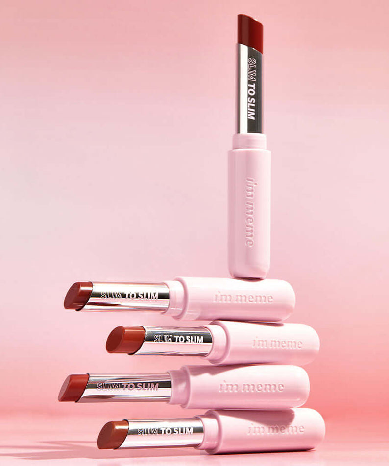 Stack of IM MEME Slim to Slim Hyaluronic Gloss Fit Lipsticks displayed on a pink background.