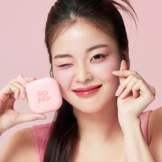 Smiling woman holding IPKN NU'EST Honey Pact 13.5g, showing compact design, against pink background. Beauty and skincare product.