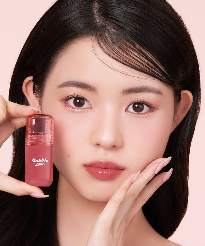 Woman holding IPKN Bubble Jam Gloss, highlighting lip gloss packaging and color on a soft pink background.