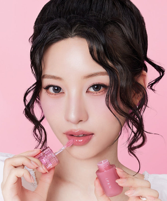 Woman applying IPKN Bubble Jam Gloss for a glossy finish on pink background