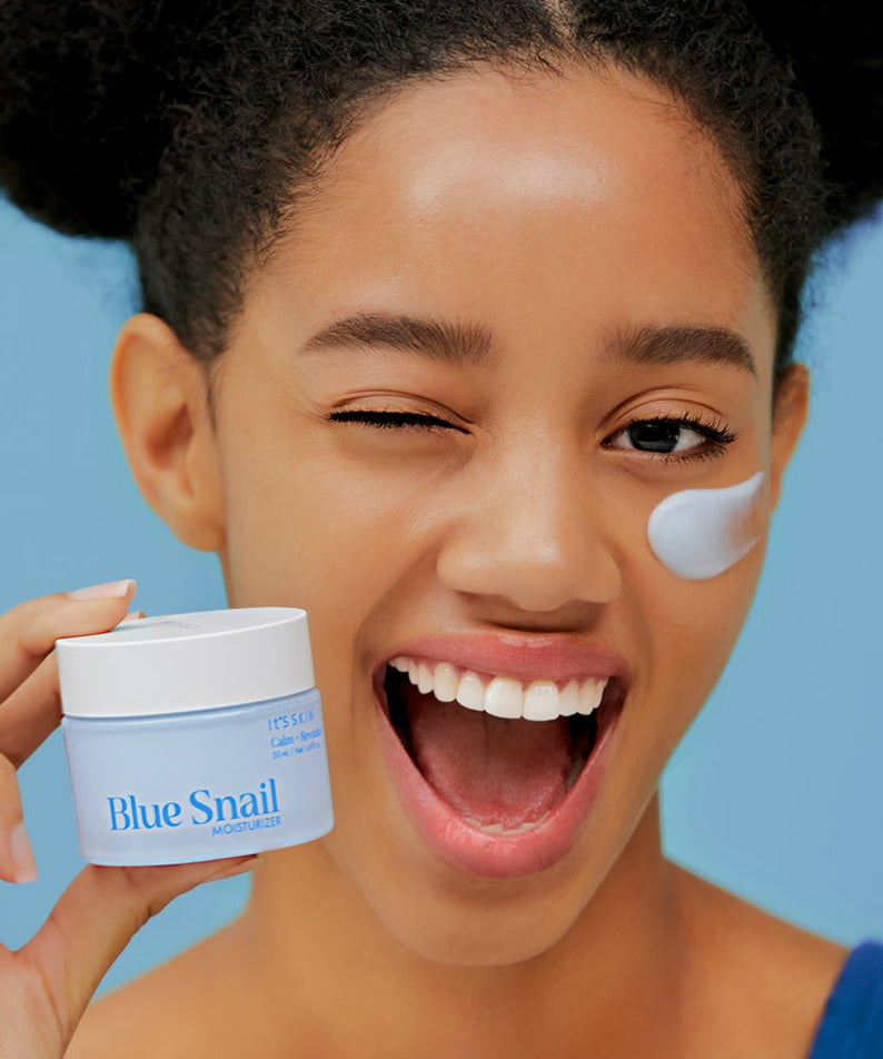 Smiling person holding IT'S SKIN Blue Snail Cream 50ml with cream on cheek, bright skin care product.
