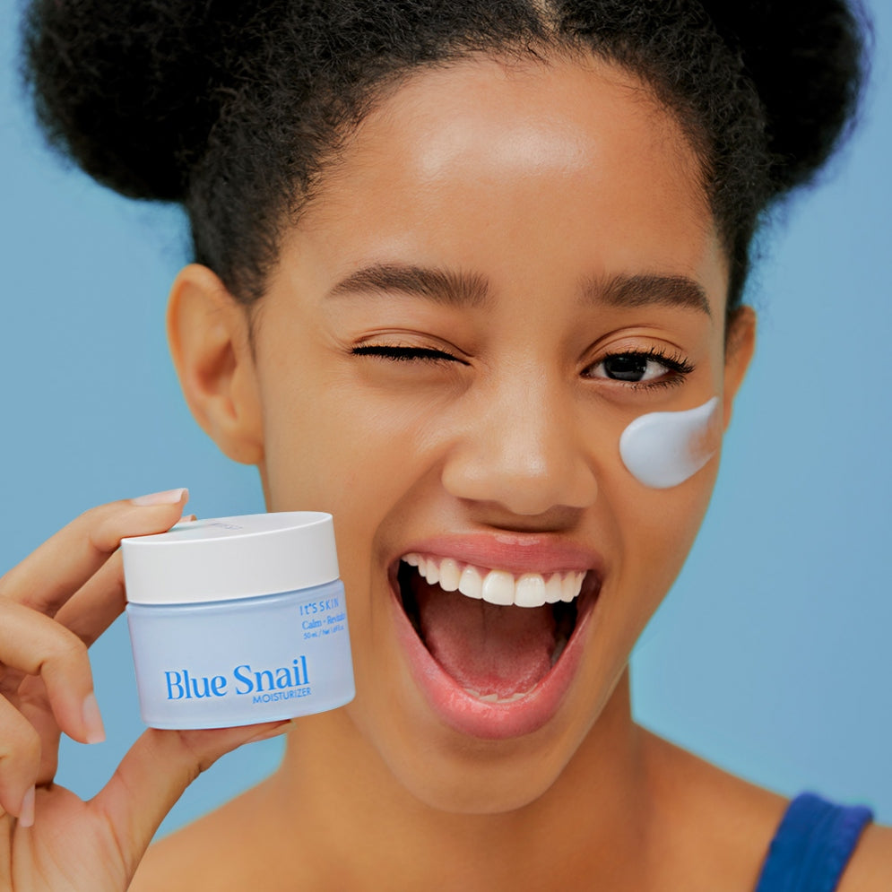 Smiling person holding IT'S SKIN Blue Snail Cream 50ml with cream on cheek, bright skin care product.