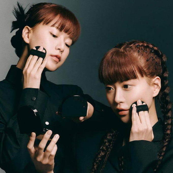 Two models applying JAVIN DE SEOUL Wink Cushion Glow 16g Â, featuring sleek black compacts against a dark background.