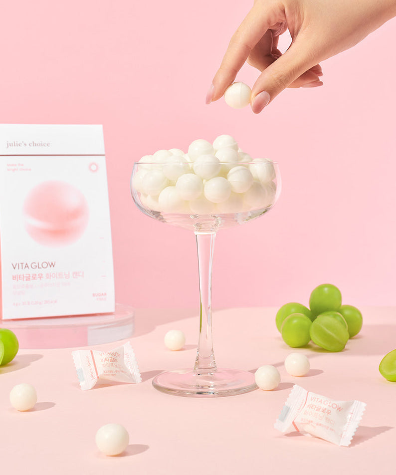 JULIE'S CHOICE Vita Glow Whitening Candy displayed in a glass with packaging and grapes on a pink background