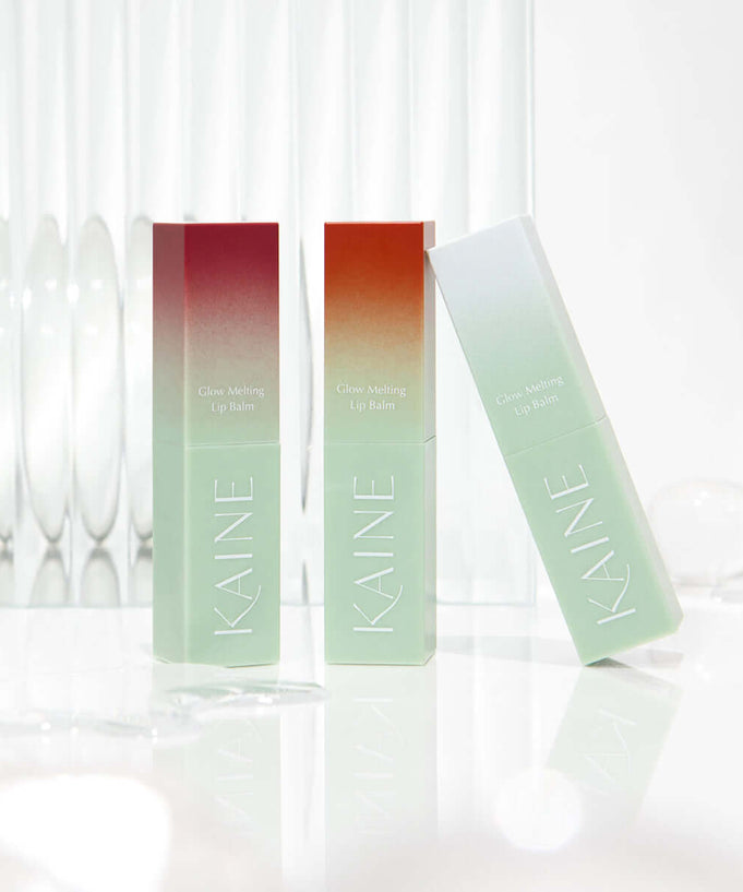 Three KAINE Glow Melting Lip Balm tubes standing on a reflective surface with a clear glass backdrop.