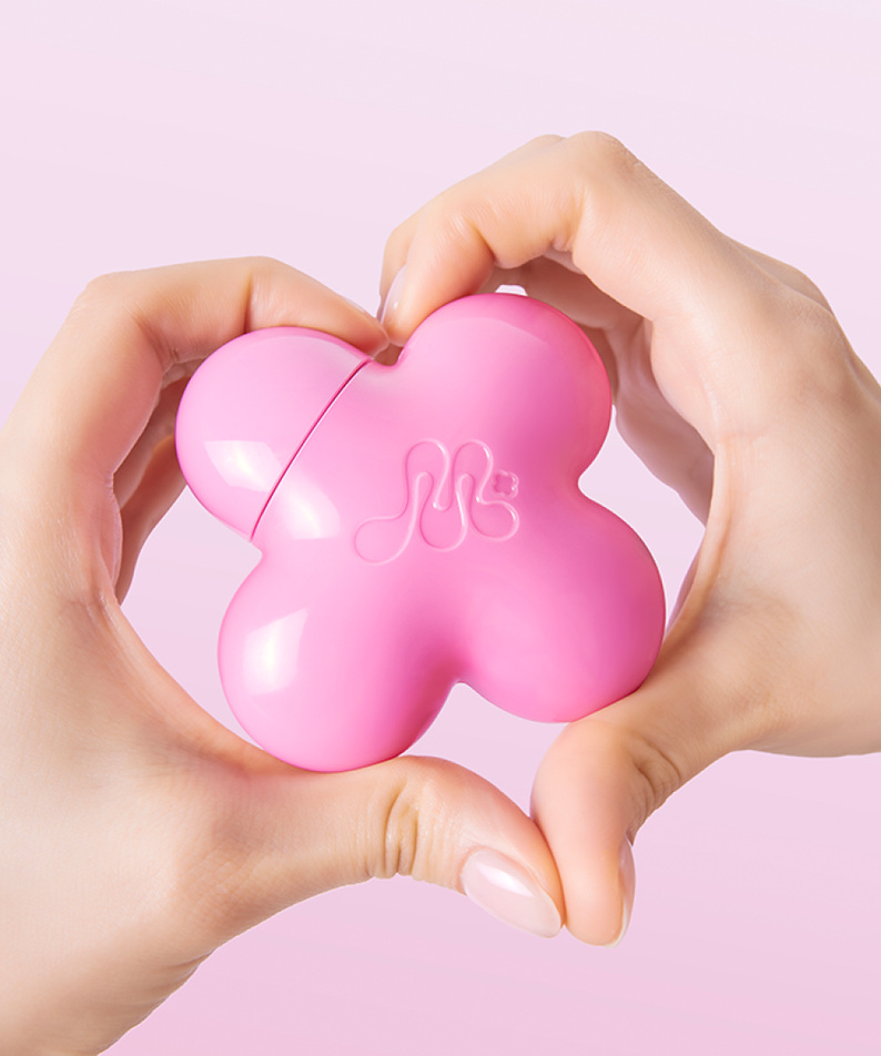 Hands holding pink clover-shaped beauty product against a pink background.