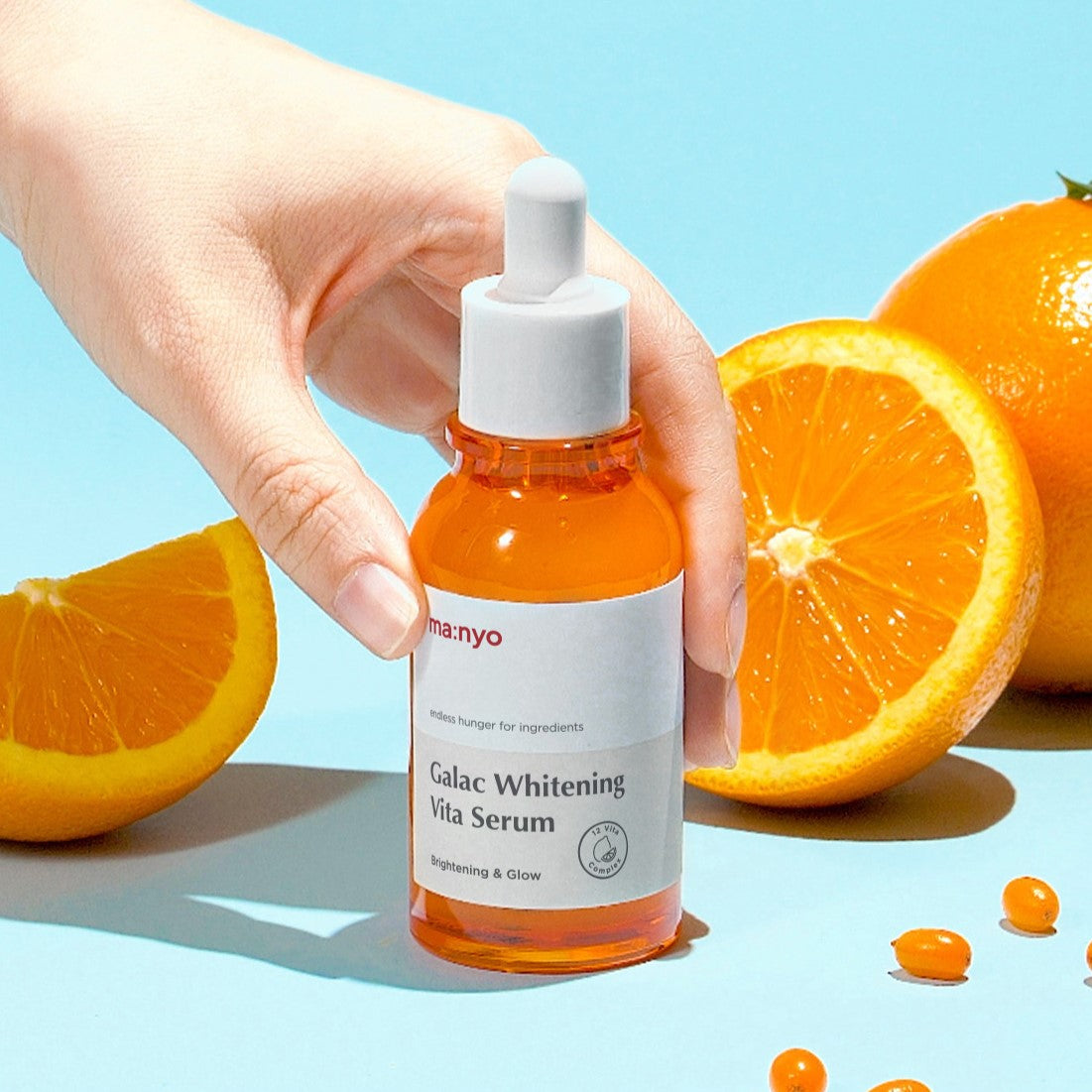 Hand holding MA:NYO Galac Whitening Vita Serum 50ml with sliced oranges in the background, showcasing the product's brightening effect.