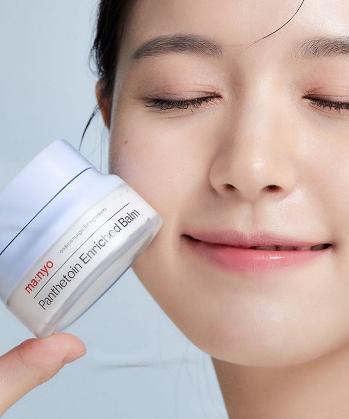 A woman applying MA:NYO Panthetoin Enriched Balm 80ml to her cheek, holding the product jar with a smile.