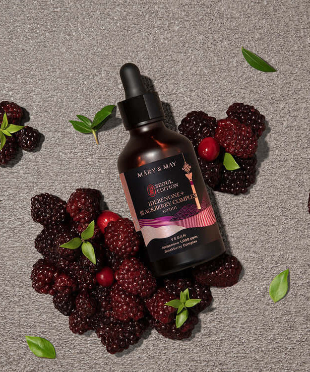 MARY & MAY Idebenone+Blackberry Complex Serum bottle surrounded by fresh blackberries on a textured surface