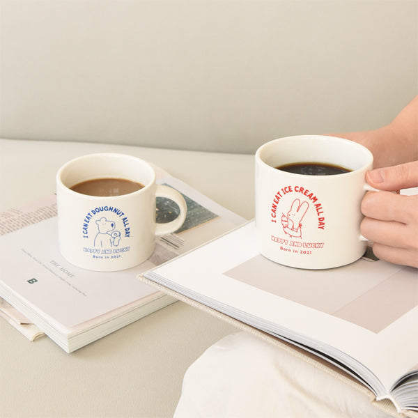 Two ceramic cups, one displaying "Enjoy Doughnut All Day" in blue and the other "I Love Ice Cream All Day" in red, filled with coffee on a book.