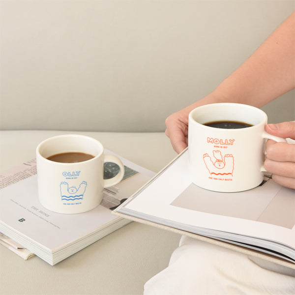 Person holding a MONOLIKE Olly Molly Mug with coffee, featuring a cute design, placed on a book; perfect for cozy moments.