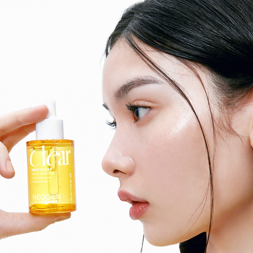Close-up of person holding NEOGEN Next Clear Spot Solution 32ml bottle near face, showcasing skincare product packaging and texture.