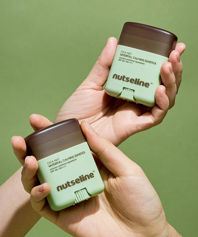 Two hands holding NUTSELINE Cica Nut Waterful Calming Sun Stick 20g against a green background.