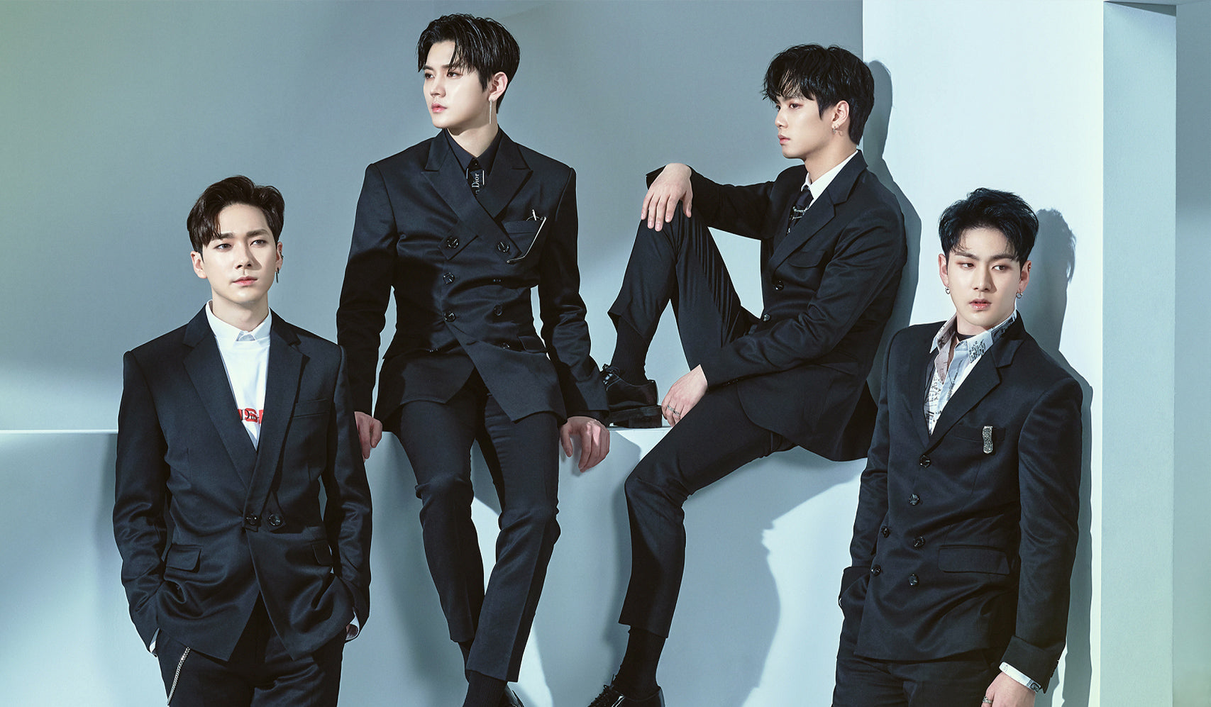 Four men in formal black suits posing with a modern background.