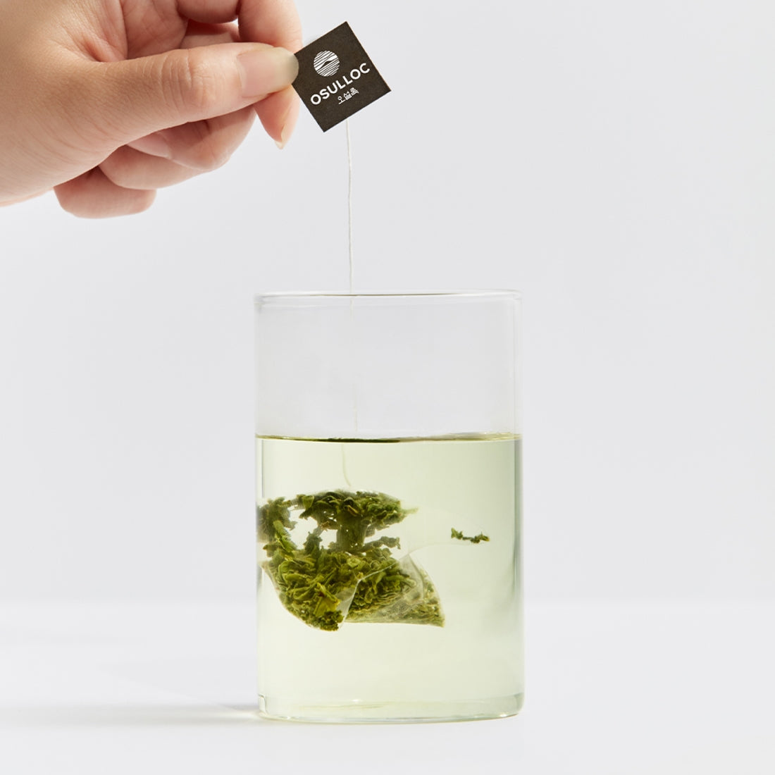Brewing OSULLOC Sejack Green Tea 20 P tea bag in a glass with fresh green leaves visible.