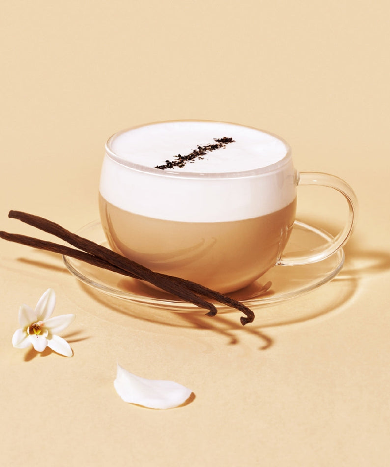 Glass cup of OSULLOC Vanilla Honey Black Tea with frothy cream, vanilla beans, and a delicate flower on a beige background.