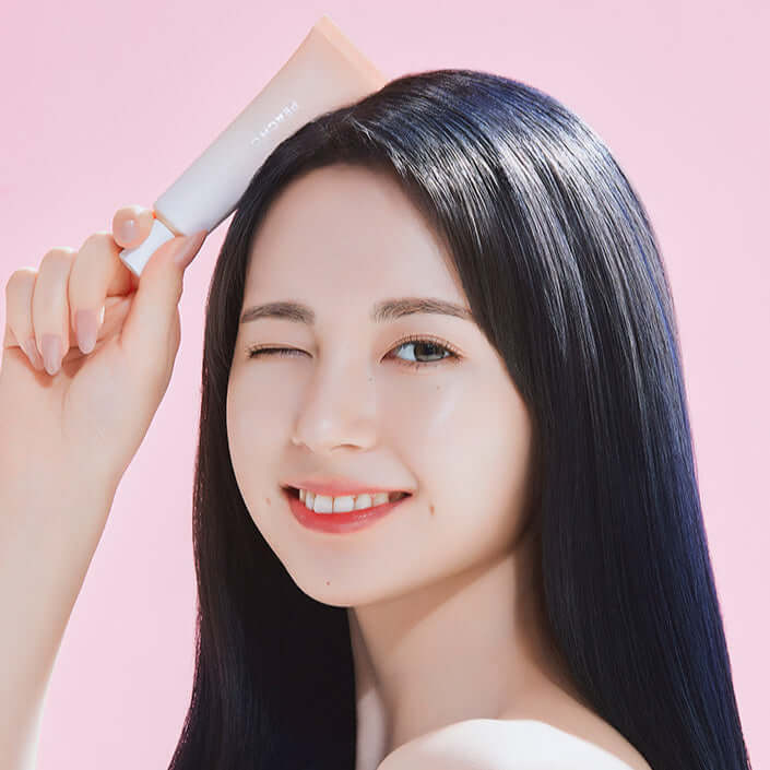 Smiling woman holding PEACH C Beige Glow Makeup Base against pink background, showcasing radiant skin effect.