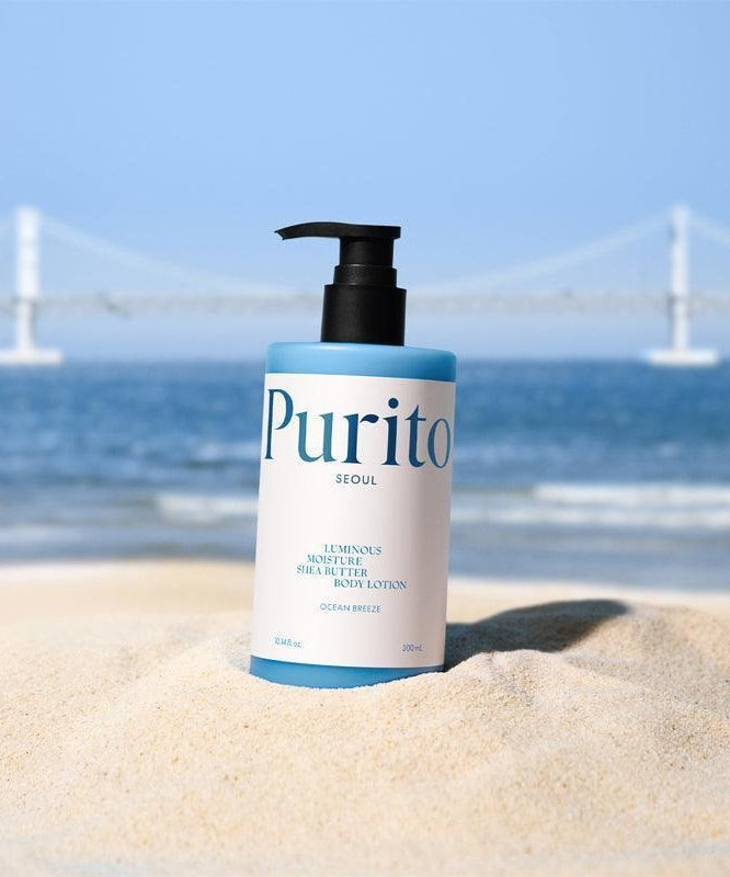 PURITO Luminous Moisture Shea Butter Body Lotion 300ml bottle on sand with a sea and bridge background