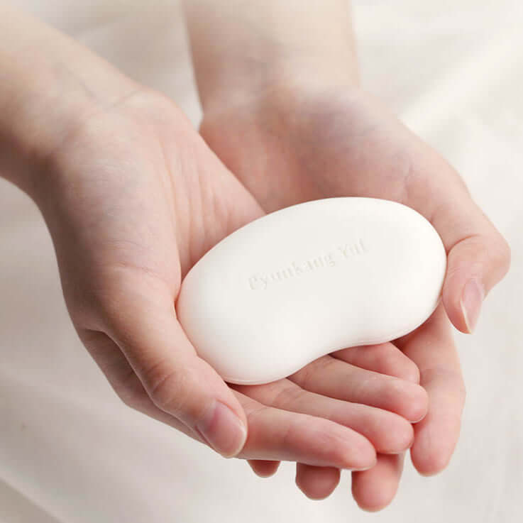 Hands holding PYUNKANG YUL Ato High Moisture Baby Soap 80g (2 pieces) on a soft fabric background.