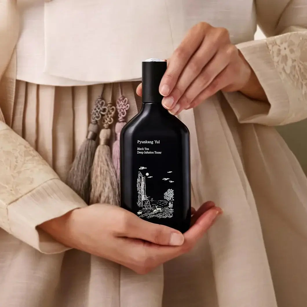 Hands holding PYUNKANG YUL Black Tea Deep Infushion Toner 130ml bottle with elegant design packaging.