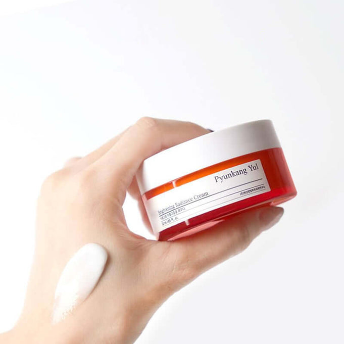 Hand holding a jar of PYUNKANG YUL Brightening Radiance Cream 50ml with cream applied on the skin, showcasing product texture.