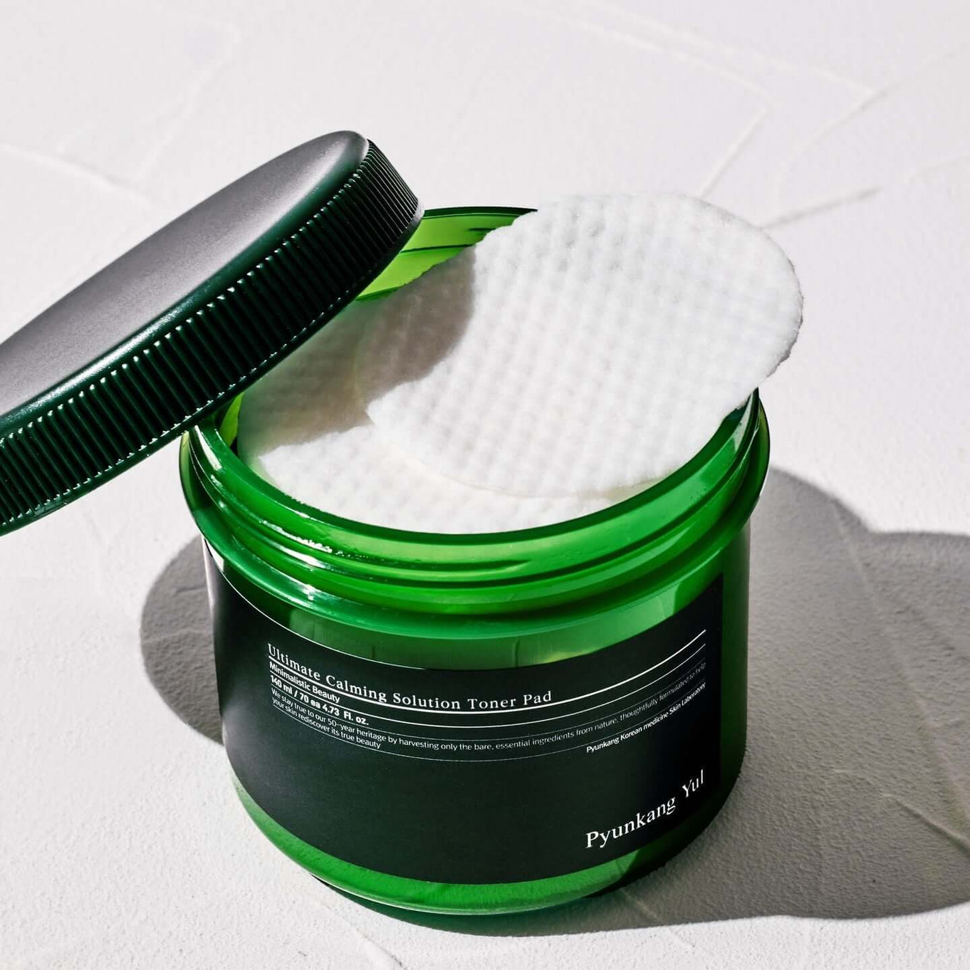 Open jar of PYUNKANG YUL Ultimate Soothing Solution Toner Pads 70 sheets 140ml with white pad halfway out on white background