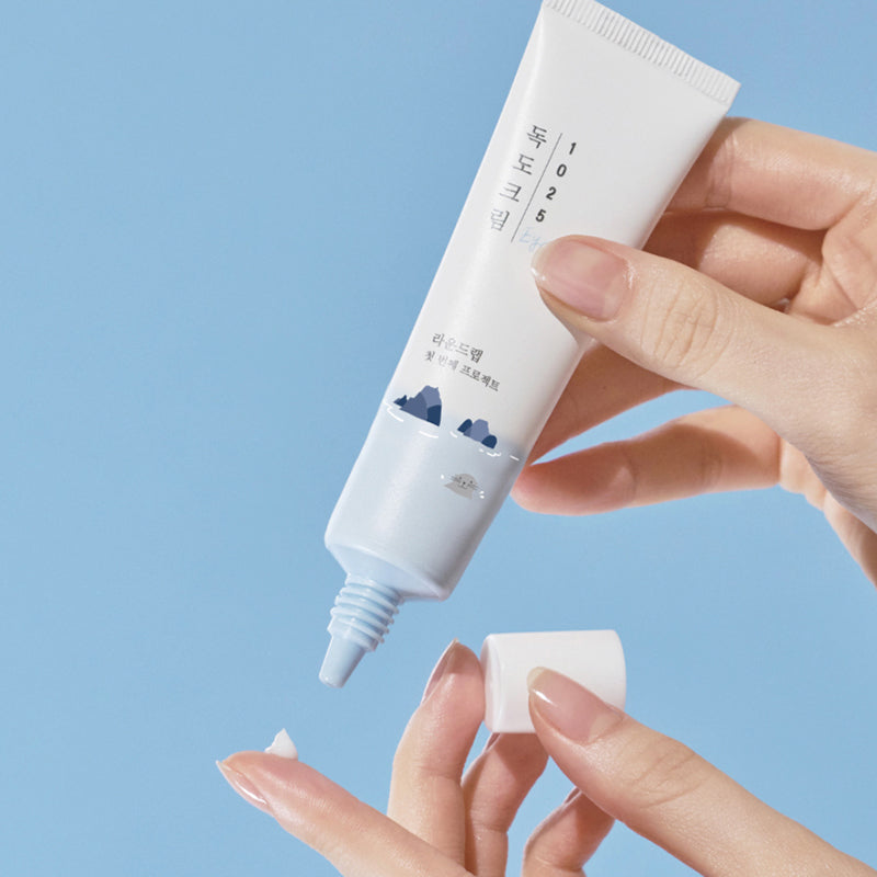 Person applying ROUND LAB 1025 Dokdo Eye Cream 30ml on finger from tube against blue background