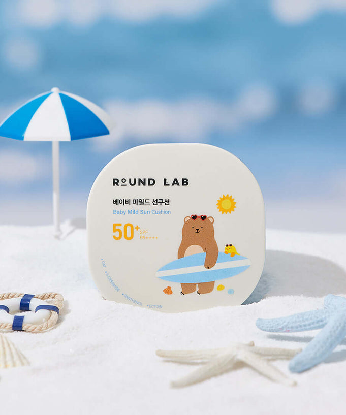 ROUND LAB Baby Mild Sun Cushion 16g with SPF 50 packaging displayed on sandy beach with small umbrella and beach toys.