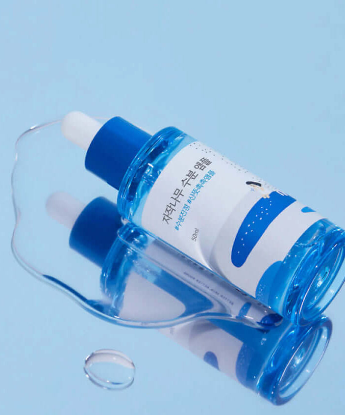 Bottle of ROUND LAB Birch Moisture Ampoule 50ml lying on a blue surface with droplets.
