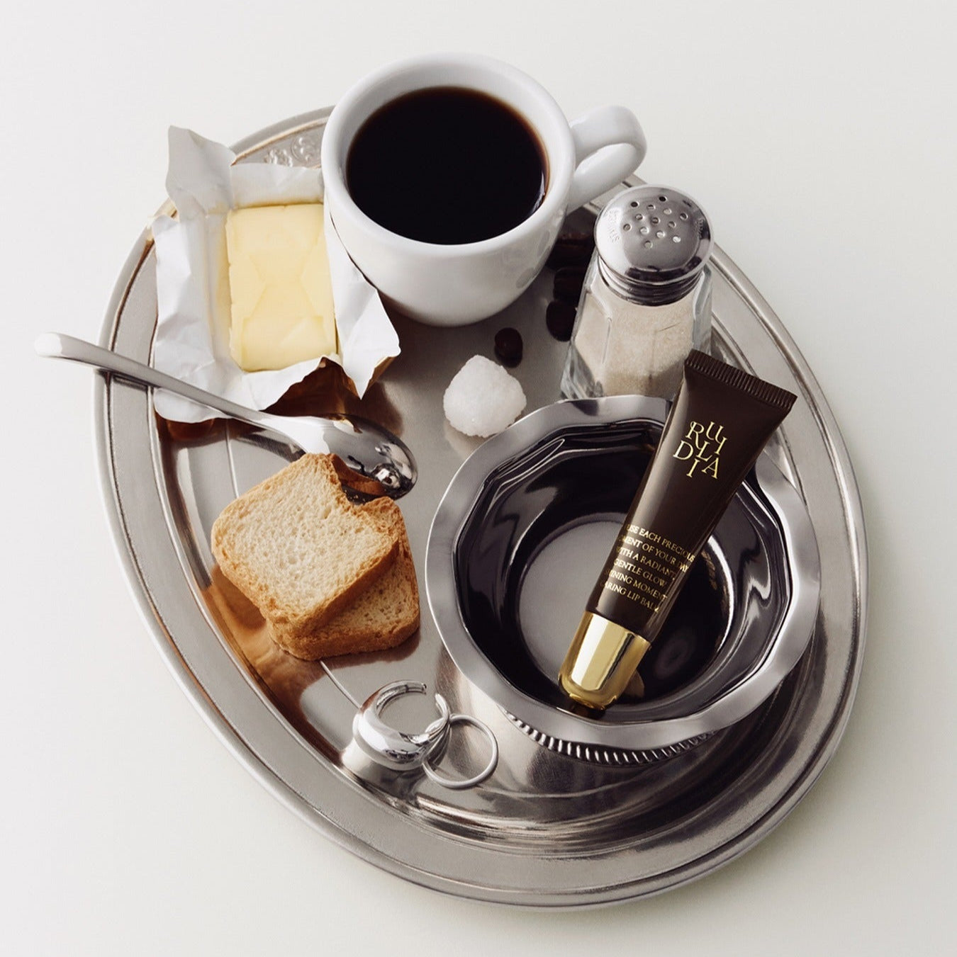 RULIDIA Shining Moment Caring Lip Balm 8g on a silver tray with coffee, toast, and butter.